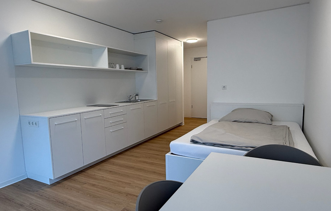 Single-Apartment 1.1 im RW63: Wohnraum mit weißer Küchenzeile, Bett und Essplatz
