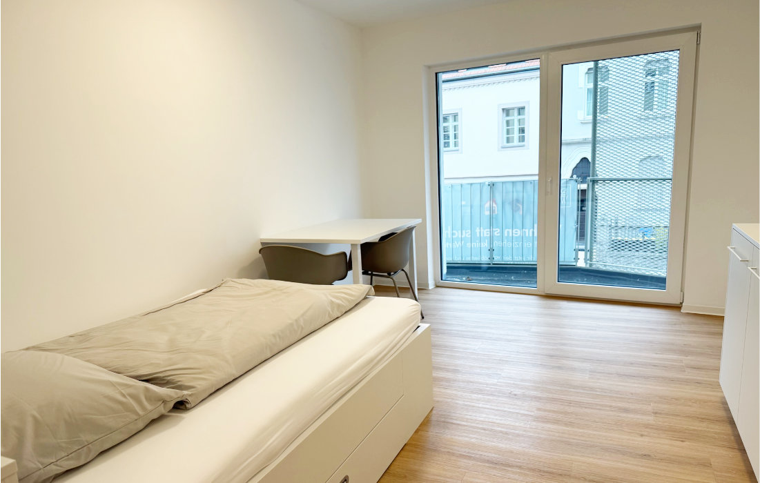 Bett, Schreibtisch mit zwei Stühlen und bodentiefe Balkontür im Single-Apartment 1.1 zur Straßenseite